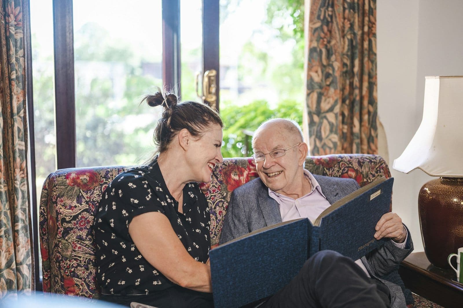 A support worker and an older man laughing together over a photo album in a front room
